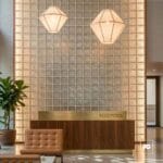Vidrio block 19x19x8-transparente-lattice-foncer-plomeria garcia-2-ok | Plomería García Hotel reception desk in front of a glass-block wall with two geometric pendant lights overhead, and a potted plant nearby.