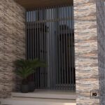 Fachaleta oxido-30x60-everest-cenezzia-2-ok | Plomería García Modern entryway with stacked stone walls, a vertical metal slat gate, and two potted plants on steps.